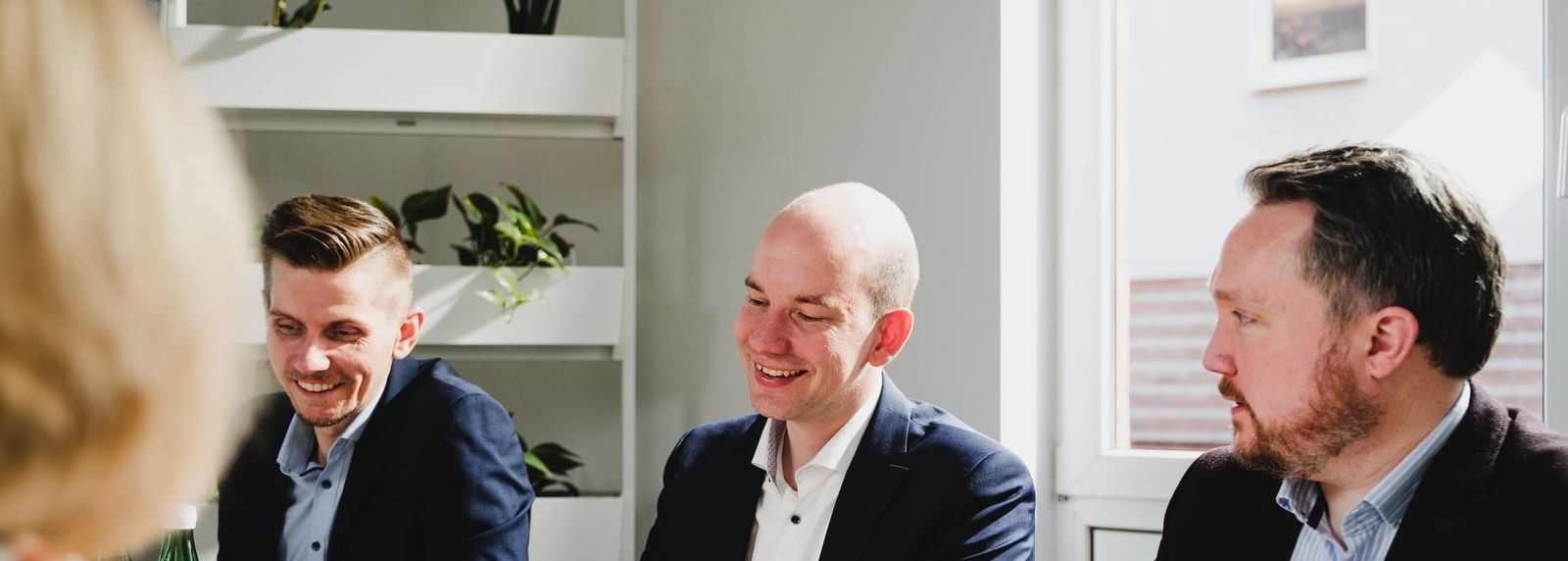 Drei Männer sitzen in einem hellen Büro im Gespräch, lachen und diskutieren miteinander; im Hintergrund Regale und Pflanzen.