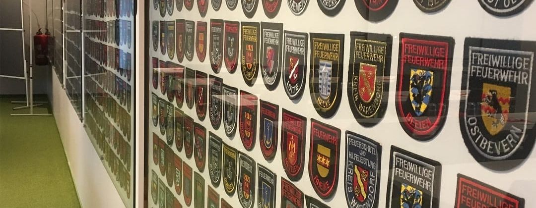 Wand mit zahlreichen bestickten Ärmelabzeichen verschiedener Freiwilliger Feuerwehren aus unterschiedlichen Städten und Gemeinden, hinter Glas ausgestellt