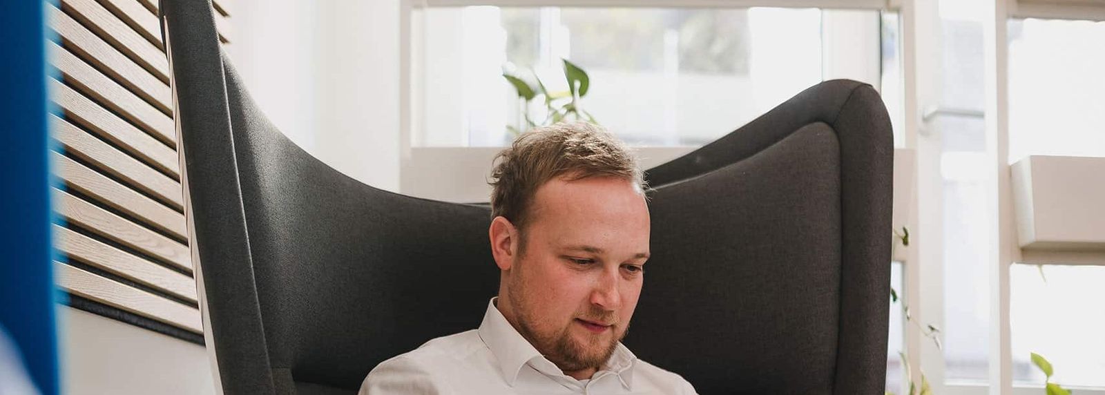 Ein Mann im weißen Hemd sitzt entspannt in einem Sessel in einem hellen Büro und arbeitet an einem Laptop; im Vordergrund ein Gesprächspartner.