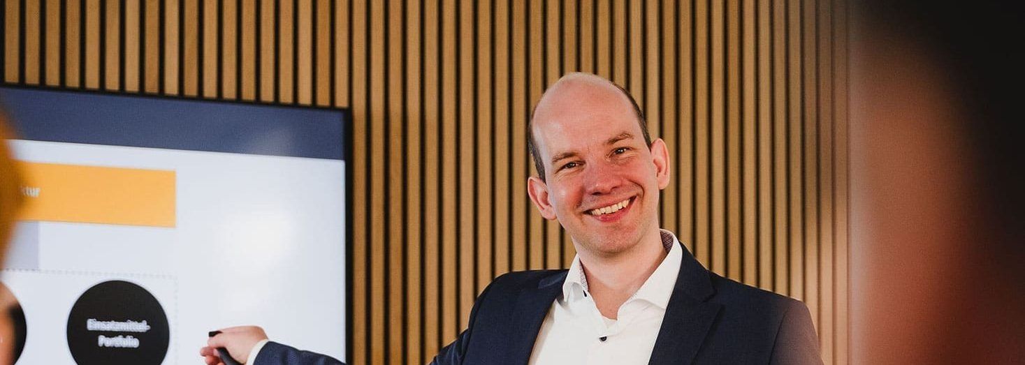 Ein lächelnder Mann im dunklen Sakko steht vor einem Bildschirm und zeigt auf eine Präsentationsfolie zur Standortplanung; im Vordergrund sitzen Zuhörende.