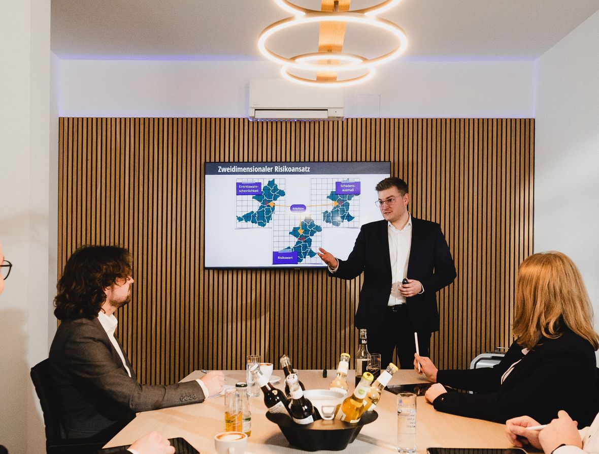 Berater erläutert im Meetingraum anhand einer Bildschirmpräsentation den zweidimensionalen Risikoansatz (Eintrittswahrscheinlichkeit, Schadensausmaß, Risikowert) für die Bedarfsplanung