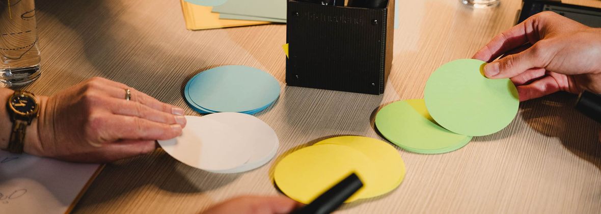 Workshop-Situation: Hände sortieren farbige Moderationskarten auf einem Tisch, auf einer gelben Karte steht handschriftlich N-KTW