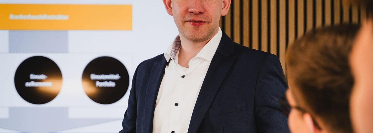 Ein Mann im dunklen Sakko präsentiert vor einem Bildschirm eine Folie zur Standortgutachten-Portfolioanalyse und gestikuliert zu zwei Zuhörern am Tisch.