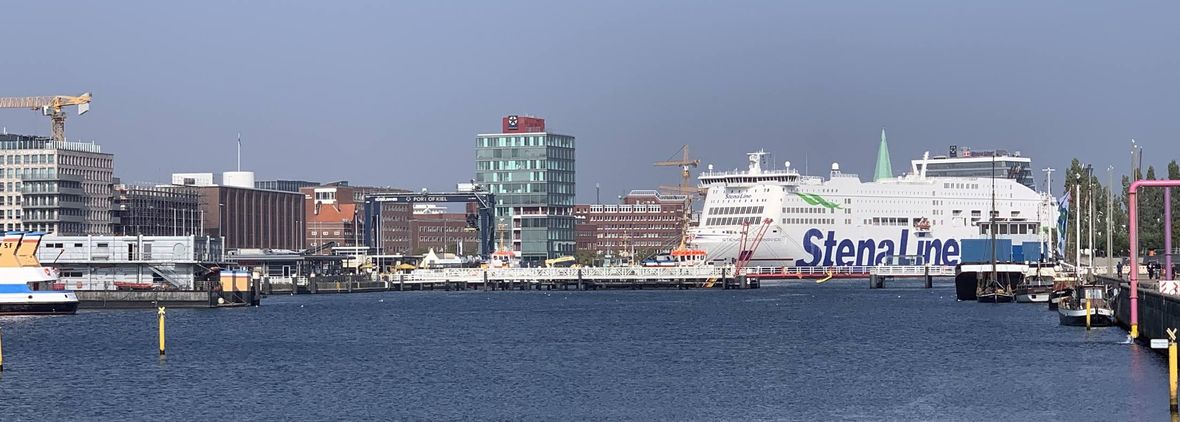 Blick über das Wasser der Hörn im Kieler Hafen. Am Ufer stehen Bürogebäude, ein gläsernes Hochhaus und Baukräne. Rechts liegt eine große weiße Fähre am Anleger, dahinter ragt eine grüne Kirchturmspitze auf.