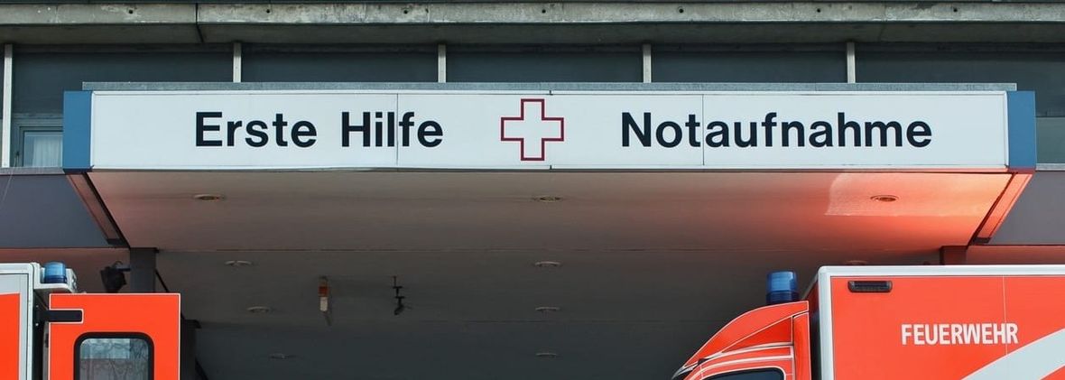 Liegendanfahrt eines Krankenhauses. Über der Zufahrt hängt ein beleuchtetes Schild mit der Aufschrift „Erste Hilfe – Notaufnahme", darunter stehen zwei Rettungswagen der Feuerwehr unter dem Vordach.