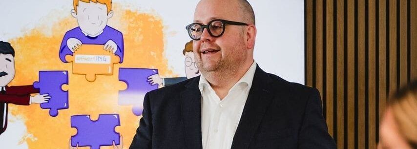Ein Mann im dunklen Anzug spricht vor einer Gruppe; hinter ihm zeigt ein Bildschirm eine Illustration, in der Figuren gemeinsam Puzzleteile mit dem antwortING-Logo zusammensetzen.