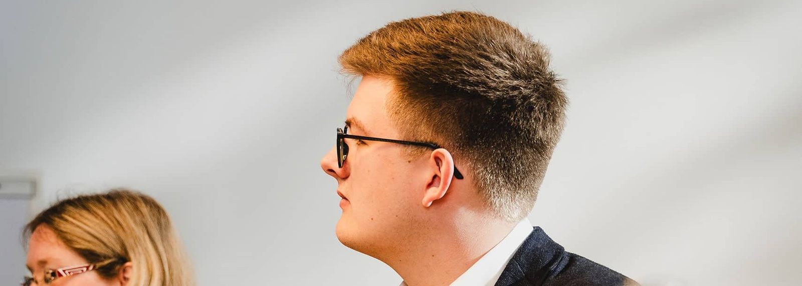 Eine Frau mit Brille und ein jüngerer Mann mit Brille im dunklen Sakko sitzen nebeneinander an einem Besprechungstisch, hören aufmerksam zu und machen sich Notizen.