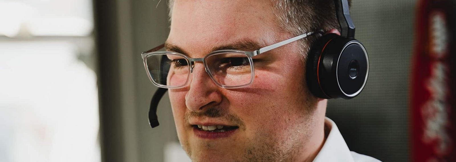 Ein junger Mann mit Brille und Headset im weißen Hemd telefoniert in einem Büro und blickt lächelnd zur Seite.
