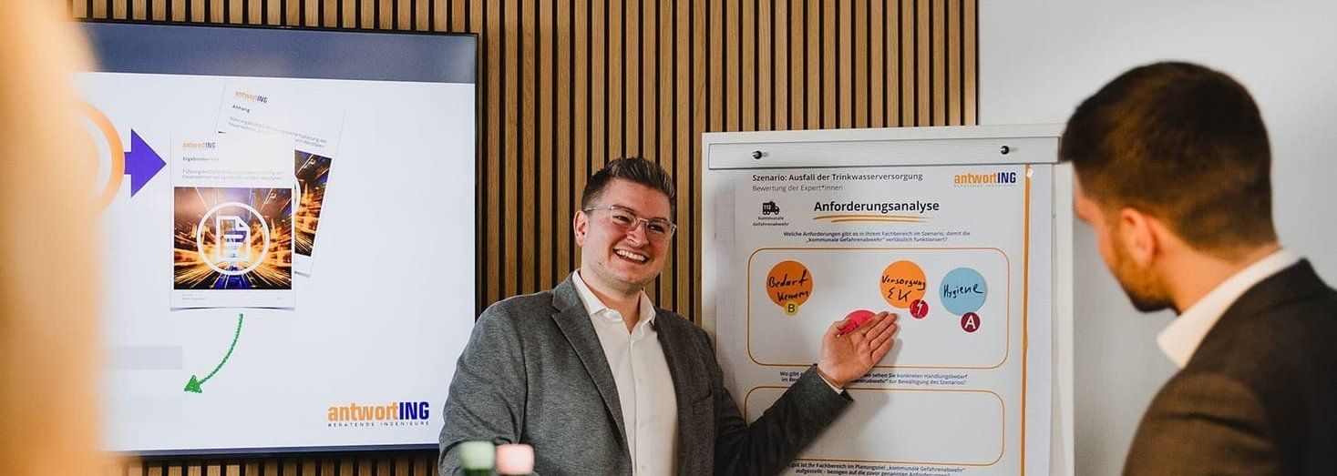 Ein lächelnder Mann präsentiert in einem Besprechungsraum an einem Flipchart eine Anforderungsanalyse; Kolleginnen und Kollegen sitzen am Tisch.