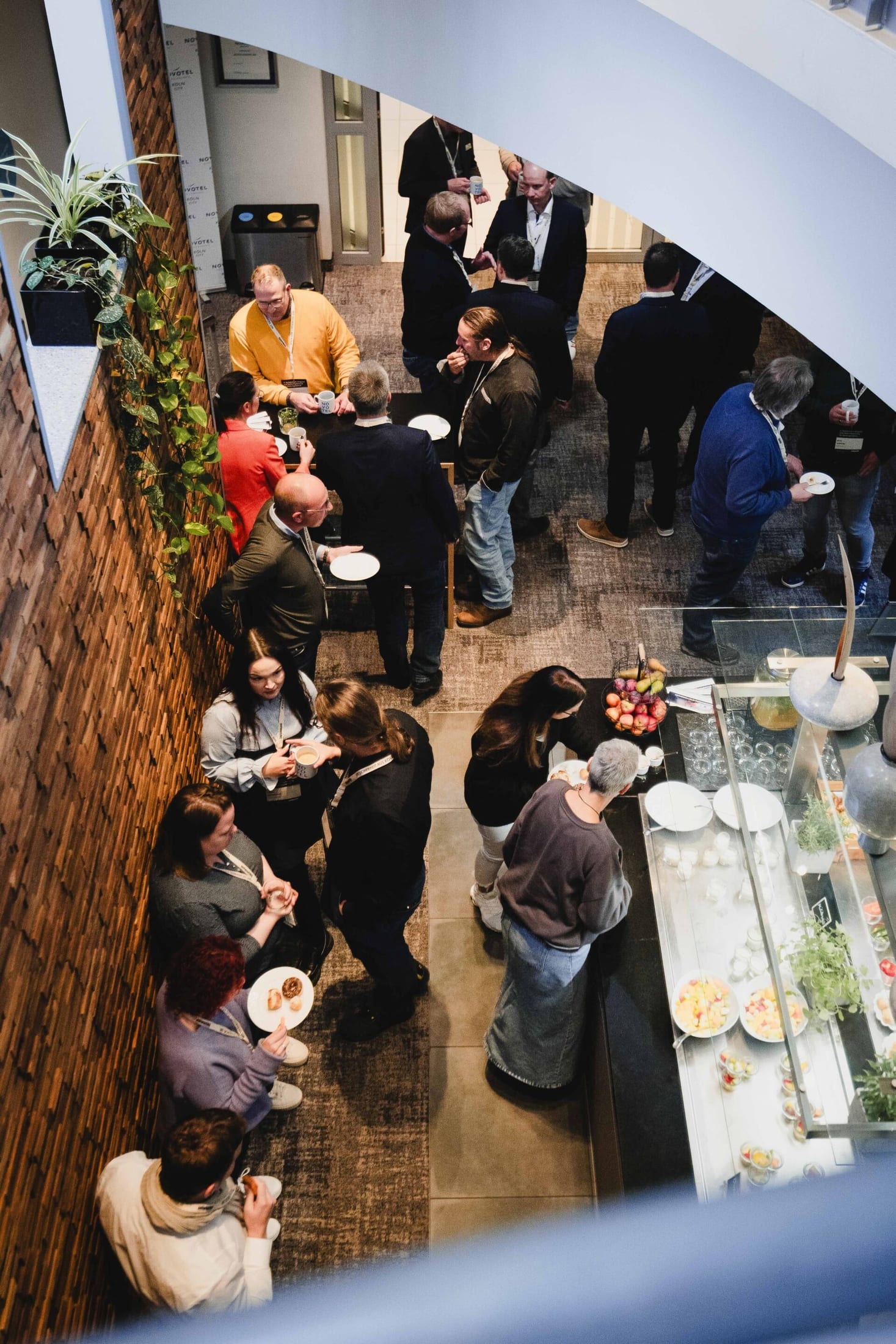 Blick von oben auf Teilnehmende des Symposiums, die in einer Pause in Grüppchen an einer Backsteinwand und einem Buffet mit Speisen zusammenstehen.
