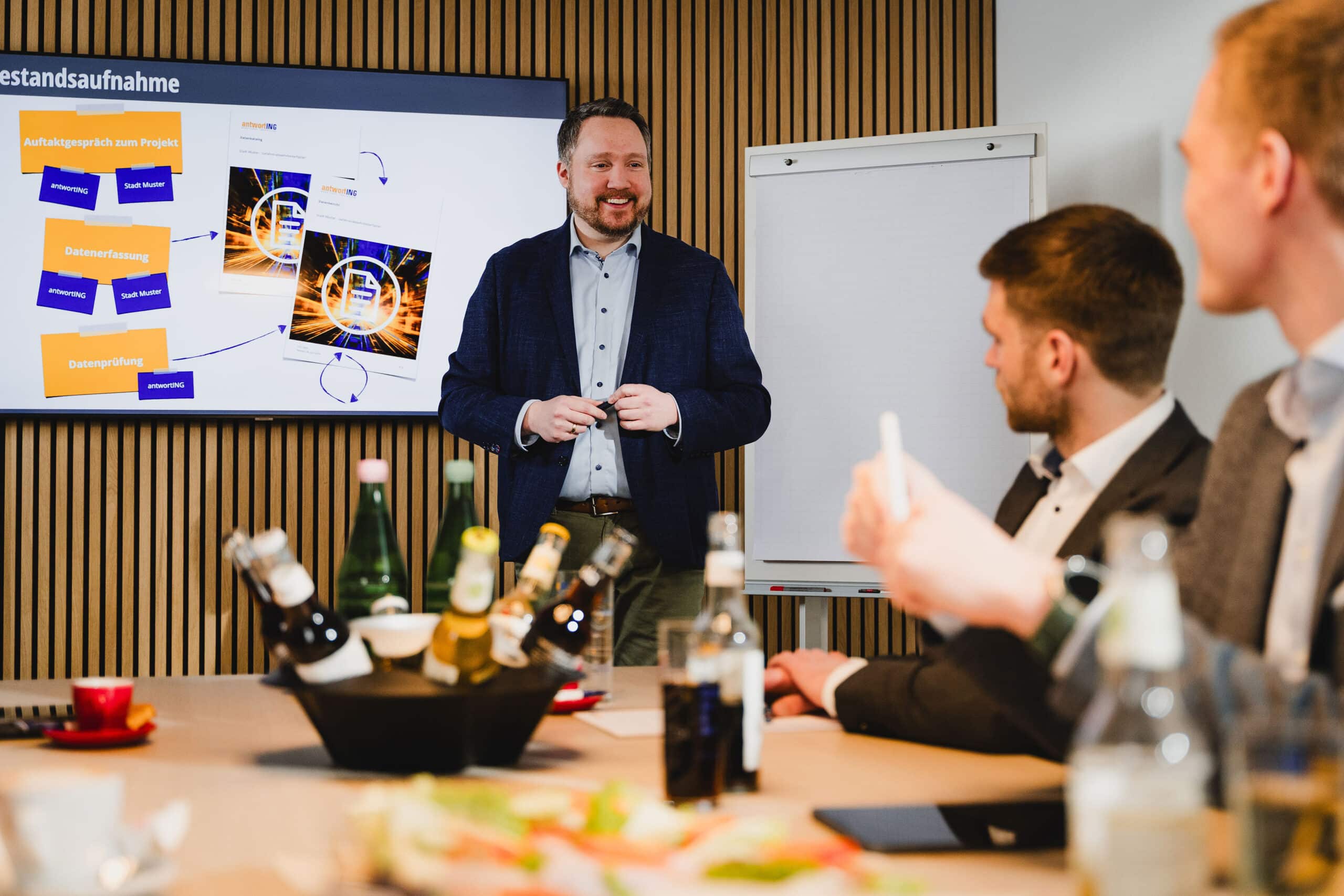 Ein Berater von antwortING moderiert einen Workshop; auf dem Bildschirm die Projektschritte Bestandsaufnahme, Datenerfassung und Datenprüfung, Kolleginnen und Kollegen arbeiten mit.