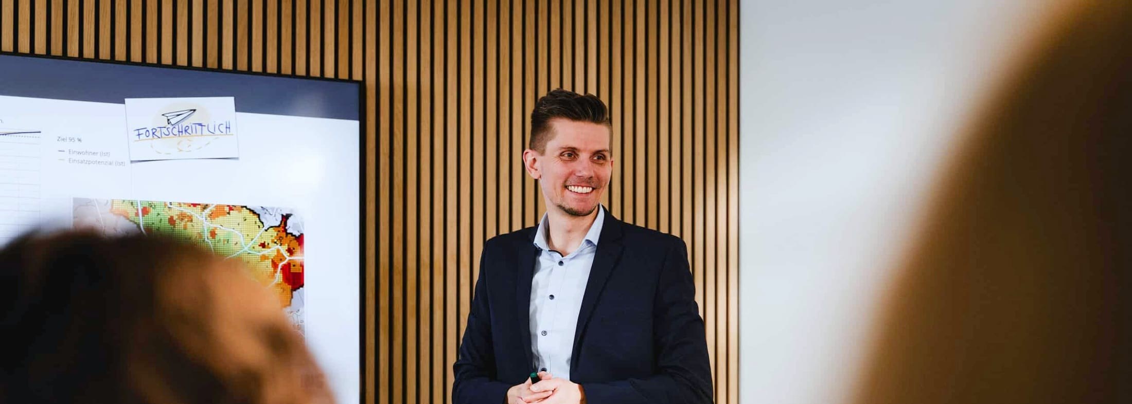 Niclas Fürst stellt im Besprechungsraum eine Erreichbarkeitsanalyse vor. Auf dem Bildschirm hinter ihm liegt eine Karte mit farbig abgestuften Fahrzeitflächen über dem Straßennetz, daneben ein Diagramm mit der Ziellinie 95 Prozent.